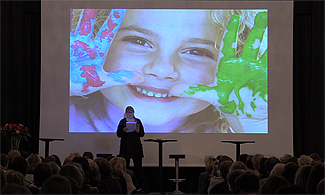 Bild på konferensscenen och en presentationsbild i bakgrunden på en flicka som visar sina målade händer.